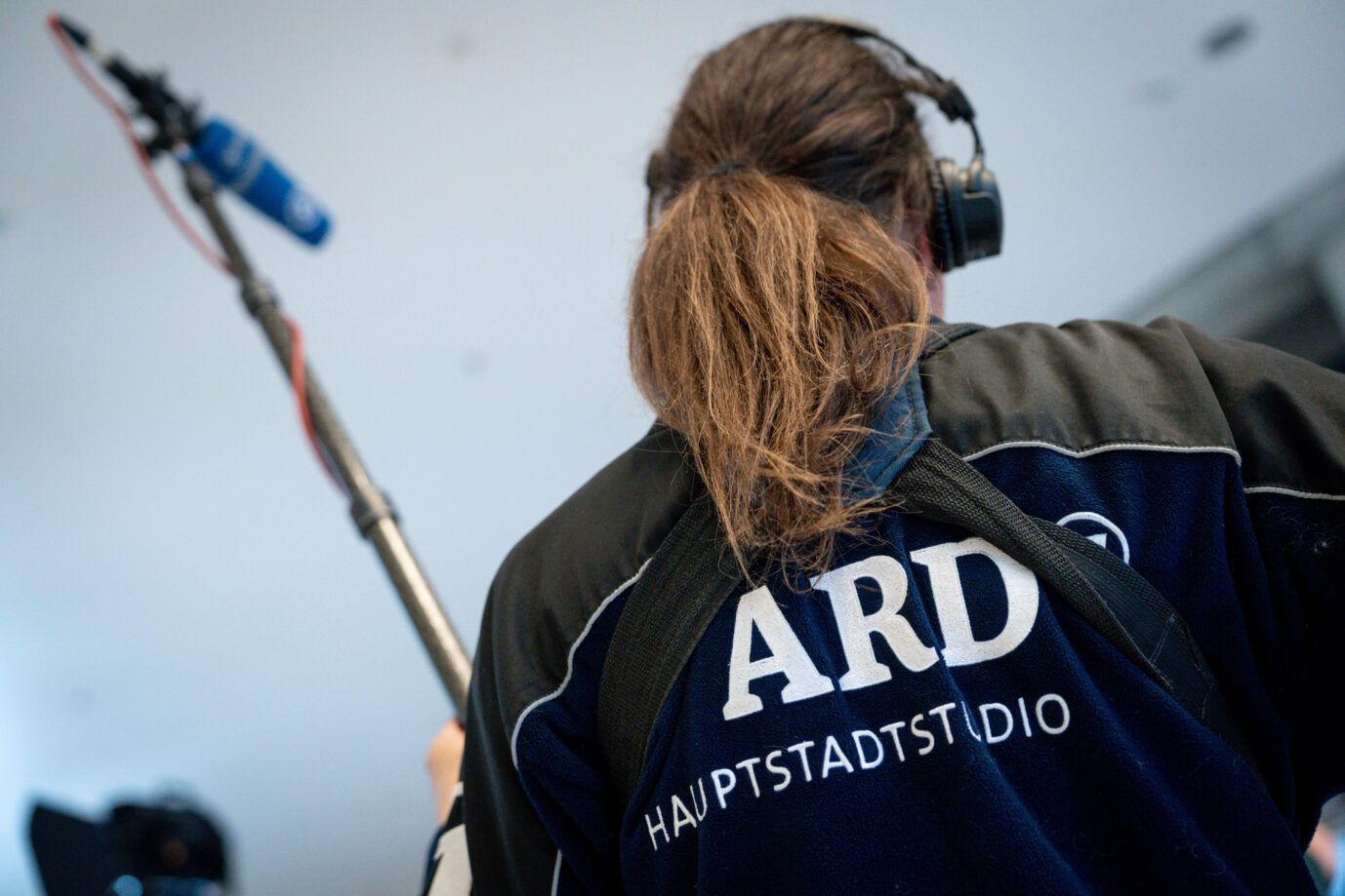 Rückansicht einer Kamerafrau oder Tontechnikerin des ARD-Hauptstadtstudios mit Mikrofonangel. Auf der Jacke ist gut sichtbar das Logo „ARD Hauptstadtstudio“ aufgedruckt. Das Bild zeigt eine Szene aus dem Arbeitsalltag der öffentlich-rechtlichen Rundfunkproduktion. Kamerafrau des ARD-Hauptstadtstudios: Zahlreiche Prominente monieren fehlende Meinungsvielfalt. Foto: picture alliance / Flashpic | Jens Krick