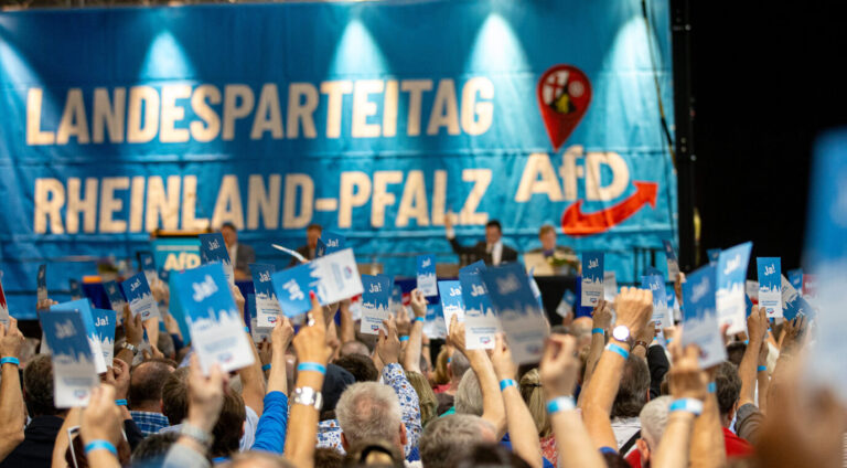 Teilnehmer eines AfD-Landesparteitags in Rheinland-Pfalz heben blaue Stimmkarten mit der Aufschrift „Ja!“ in die Höhe. Im Hintergrund ist eine große blaue Bühnenkulisse mit der Aufschrift „Landesparteitag Rheinland-Pfalz AfD“ sowie einem roten Pfeil und Landeswappen zu sehen. Auf dem Podium sitzen mehrere Personen hinter Mikrofonen. Delegierte stimmen auf dem Landesparteitag der AfD in Idar-Oberstein ab. Beim Parteitag stellt die AfD ihre Liste für die Landtagswahlen auf. Gute Laune: AfD-Mitglieder in Rheinland-Pfalz dürfen weiterhin in den Staatsdienst. Foto: picture alliance/dpa | Laszlo Pinter