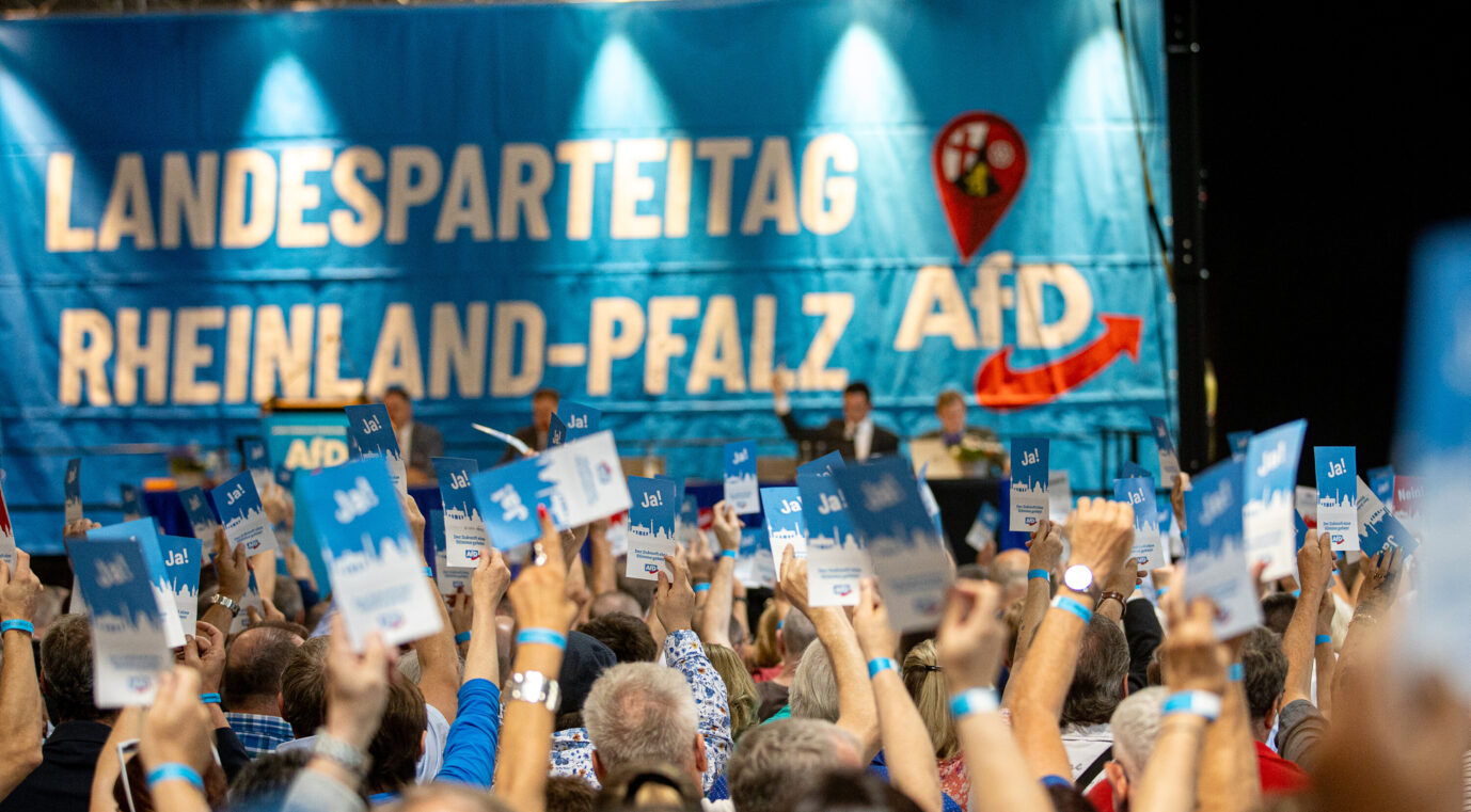 Teilnehmer eines AfD-Landesparteitags in Rheinland-Pfalz heben blaue Stimmkarten mit der Aufschrift „Ja!“ in die Höhe. Im Hintergrund ist eine große blaue Bühnenkulisse mit der Aufschrift „Landesparteitag Rheinland-Pfalz AfD“ sowie einem roten Pfeil und Landeswappen zu sehen. Auf dem Podium sitzen mehrere Personen hinter Mikrofonen. Delegierte stimmen auf dem Landesparteitag der AfD in Idar-Oberstein ab. Beim Parteitag stellt die AfD ihre Liste für die Landtagswahlen auf. Gute Laune: AfD-Mitglieder in Rheinland-Pfalz dürfen weiterhin in den Staatsdienst. Foto: picture alliance/dpa | Laszlo Pinter