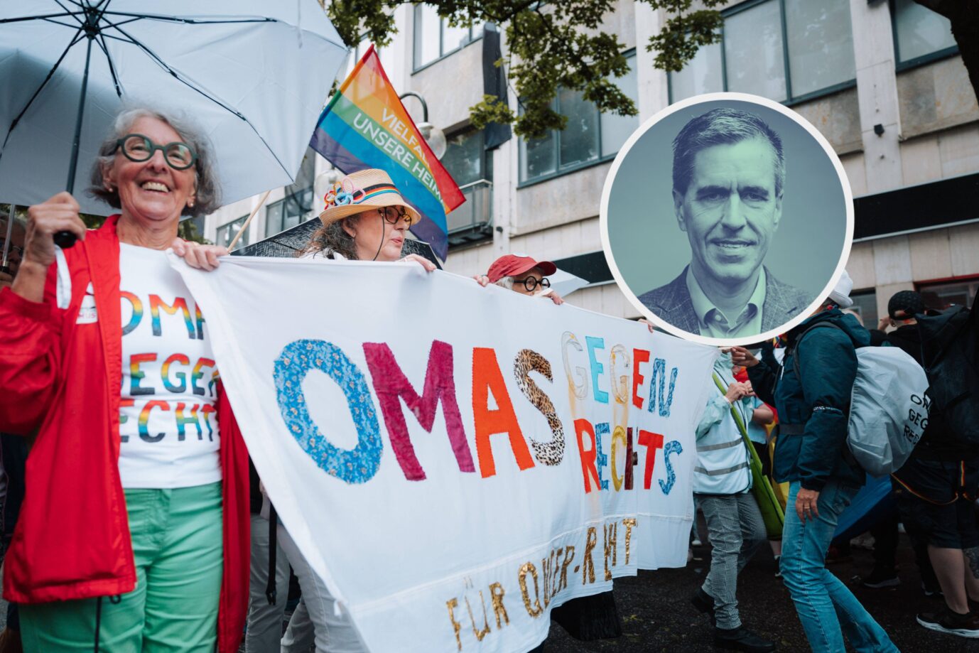 Demonstrantinnen des Bündnisses „Omas gegen Rechts“ halten bei einer Kundgebung ein selbstgebasteltes Banner mit regenbogenfarbenem Schriftzug. Im Hintergrund ist eine Regenbogenfahne mit der Aufschrift „Vielfalt unsere Heimat“ zu sehen. In das Bild montiert ist ein Porträtfoto eines Mannes, das stilisiert und kreisförmig hervorgehoben wurde – möglicherweise eine kritische oder satirische Gegenüberstellung. „Omas gegen Rechts“ in Köln: Unterstützt mit Steuergeld demonstrieren gegen Andersdenkende. Feinde der Freiheit. Foto: IMAGO / Gabor Baumgarten
