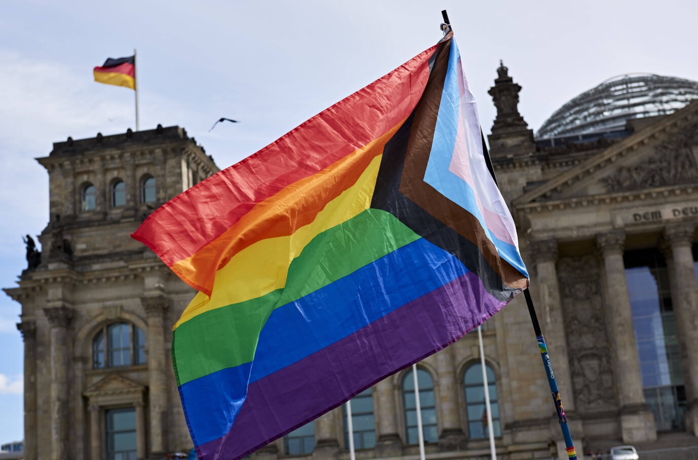 Geschlecht. Vor einem verwaschenen Himmel und dem Berliner Reichstag weht eine Regenbogen- und Transgender-Fahne im Wind – Symbolbild dafür, daß der Berliner Senat den Schutz sexueller Identitäten im Grundgesetz verankern will
