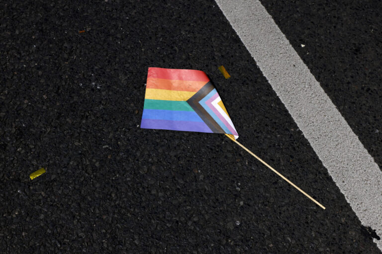 Die Flagge der Schwulen, Lesben und Transpersonen: Auf dem Asphalt einer Straße liegt eine kleine Progressive-Pride-Fahne mit abgebrochenem Holzstab. Die Szene stammt vom Christopher Street Day 2023 in Köln. Dort wurde unter dem Motto „Für Menschenrechte. Viele. Gemeinsam. Stark“ ein dreitägiges Straßenfest veranstaltet. Der ColognePride gilt nach Berlin als zweitgrößte CSD-Parade Deutschlands. Für die Parade am 9. Juli 2023 rechneten die Veranstalter mit etwa 1,5 Millionen Teilnehmern und Zuschauern entlang der Strecke. Das Foto symbolisiert den Kontrast zwischen bunter Sichtbarkeit und verletzlicher Realität sexueller Minderheiten im öffentlichen Raum. Eine Regenbogenfahne liegt auf dem Boden: Angreifer auf LSBTQ-Personen stammen meist nicht aus Deutschland. Foto: IMAGO / Panama Pictures