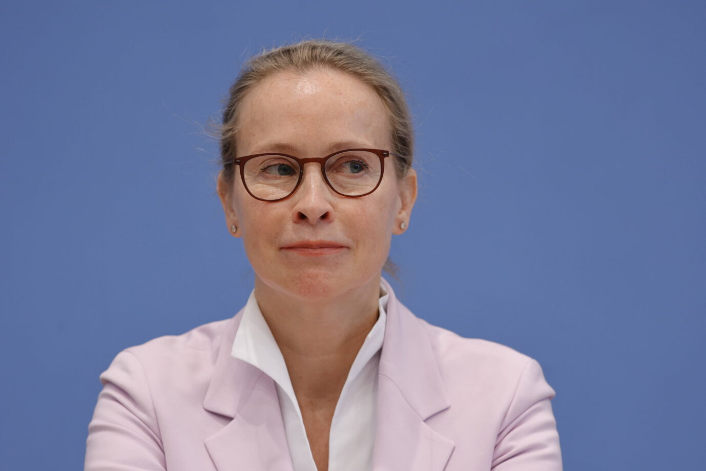Frauke Brosius-Gersdorf, Kandidatin für das Bundesverfassungsgericht, bei einer Pressekonferenz vor blauem Hintergrund. Sie trägt eine Brille und einen hellvioletten Blazer über weißem Hemd und blickt leicht zur Seite. Ihre Miene wirkt kontrolliert und nachdenklich.