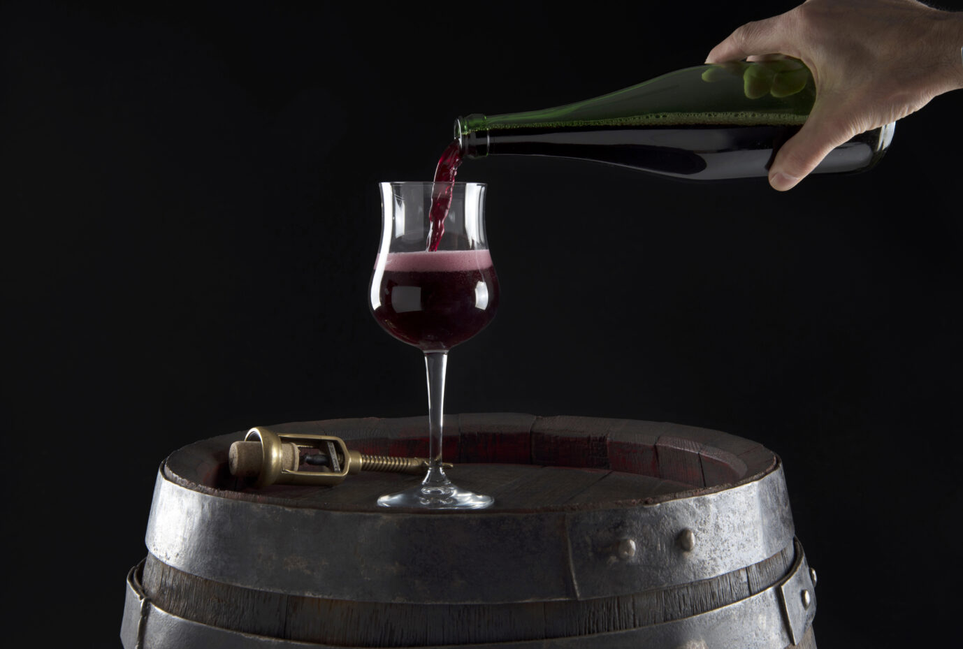 Auf einem Fass steht ein Weinglas, aus der rechten Bildhälfte kommt wiederum ein Hand und gießt frisch und fruchtig schäumenden Lambrusco in dieses Glas ein