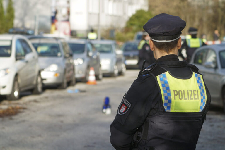 Ein uniformierter Polizist steht vor einem Parkplatz, der mit Absperrband umringt ist – ein Tatort, möglicherweise der einer Gruppenvergewaltigung
