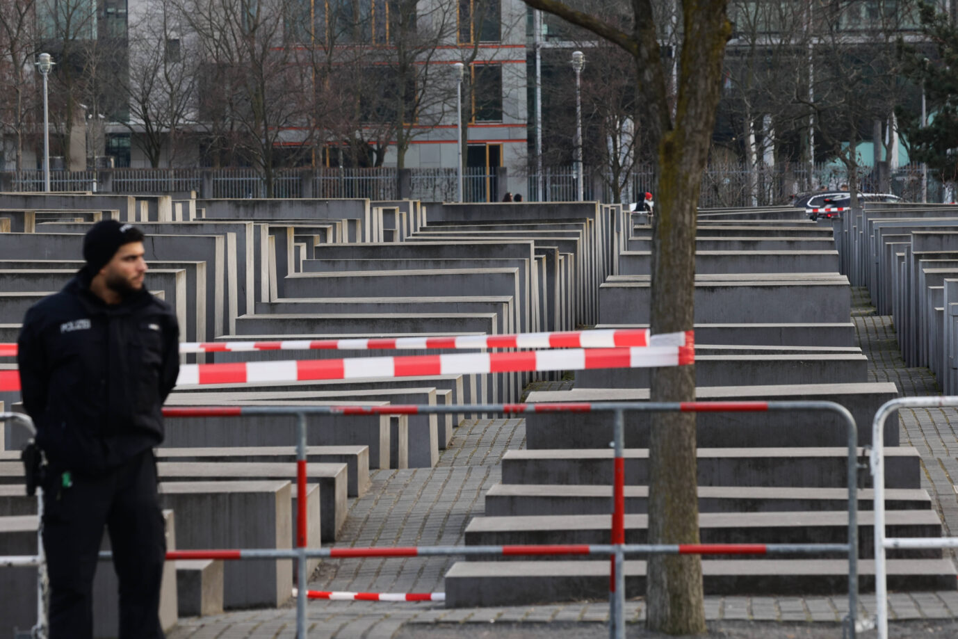 Ein Berliner Polizist steht vor dem abgesperrten Holocaust-Mahnmal in Berlin. Rote und weiße Absperrbänder sperren den Zugang zum Stelenfeld. Im Hintergrund sind die grauen Betonquader des Denkmals sowie moderne Bürogebäude zu sehen. Winterliche, kahle Bäume säumen den Ort. Das Holocaust-Mahnmal in Berlin: Hier stach der Syrer zu. Foto: IMAGO / NurPhoto
