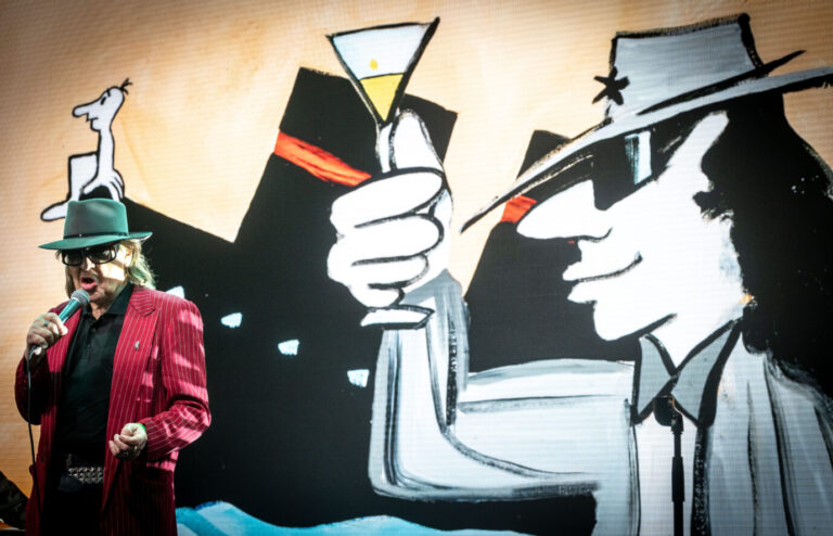 Udo Lindenberg steht mit Mikrofon und Hut auf einer Bühne, vor ihm ein großflächiges Bild seiner eigenen Kunst. Das Gemälde im Hintergrund zeigt eine cartoonhafte Figur im Anzug mit Sonnenbrille und Martini-Glas, stilisiert im typischen Lindenberg-Stil. Die Szene wirkt wie eine Verschmelzung von Performer und Kunstfigur. Das Motiv stammt aus Lindenbergs „Likörellen“-Reihe, die für ihren lakonischen Humor und politischen Unterton bekannt ist. Unter anderem gemalt mit Eierlikör. Udo Lindenberg bei der Eröffnung des „Udoversums“ im Hof des Schloß Oberhausen. Foto: IMAGO / Funke Foto Services