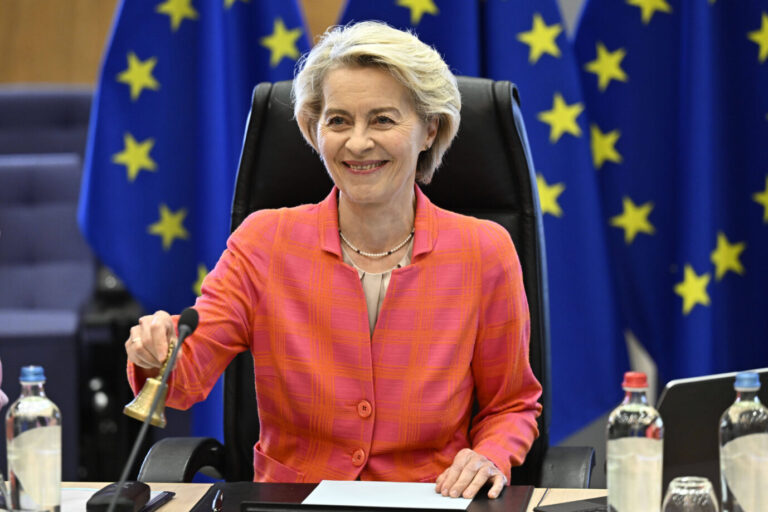 Ursula von der Leyen sitzt an einem Konferenztisch in einem Sitzungssaal, vor ihr Mikrofone, Papiere und Wasserflaschen. Sie trägt ein pink-orangekariertes Jackett, lächelt und hebt mit der rechten Hand eine kleine Glocke. Im Hintergrund stehen mehrere EU-Flaggen mit gelben Sternen auf blauem Grund. Ursula von der Leyen in Brüssel: Sie darf auch weiter die Glocke als Kommissionspräsidentin läuten. Foto: IMAGO / ZUMA Press Wire
