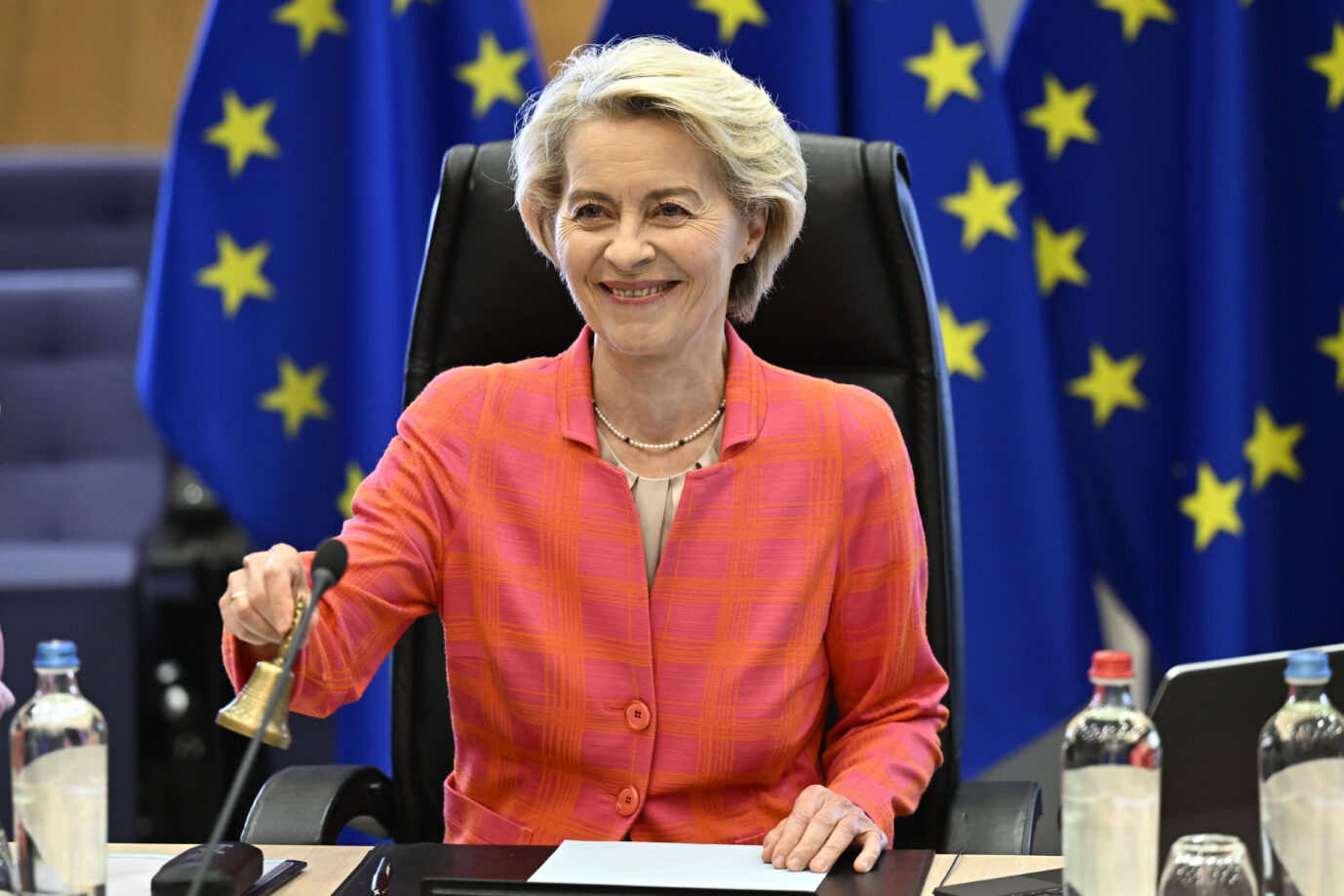 Ursula von der Leyen sitzt an einem Konferenztisch in einem Sitzungssaal, vor ihr Mikrofone, Papiere und Wasserflaschen. Sie trägt ein pink-orangekariertes Jackett, lächelt und hebt mit der rechten Hand eine kleine Glocke. Im Hintergrund stehen mehrere EU-Flaggen mit gelben Sternen auf blauem Grund. Ursula von der Leyen in Brüssel: Sie darf auch weiter die Glocke als Kommissionspräsidentin läuten. Foto: IMAGO / ZUMA Press Wire