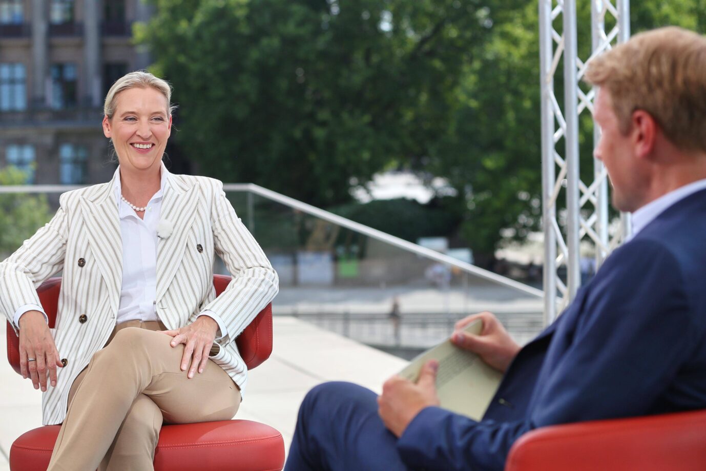 Alice Weidel sitzt beim ARD-Sommerinterview 2024 im Freien auf einem roten Stuhl und blickt lächelnd in Richtung des Interviewers Markus Preiß, der ihr gegenüber sitzt. Im Hintergrund sind Bäume, Gebäude und ein Geländer mit Glasverkleidung zu sehen. Die Szene spielt sich auf einer erhöhten Terrasse im Berliner Regierungsviertel ab. AfD-Chefin Alice Weidel im ARD-Sommerinterview: Wegen linker Störer wünscht die Partei eine Wiederholung. Foto: IMAGO / dts Nachrichtenagentur
