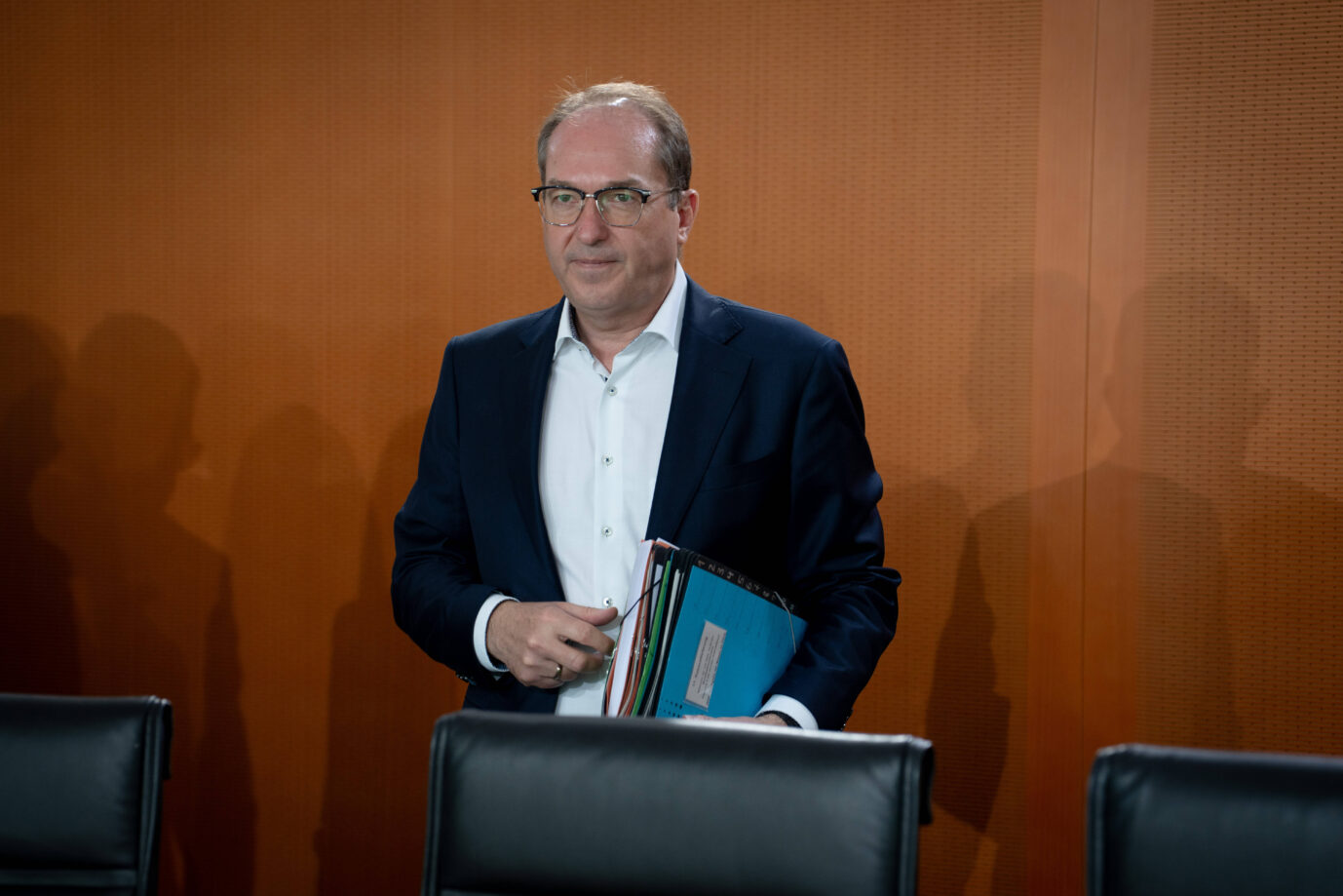 Ein Mann mittleren Alters mit Brille und Halbglatze steht in einem Konferenzraum vor einer holzvertäfelten Wand. Er trägt ein dunkles Sakko über einem weißen Hemd und hält mehrere Aktenordner unter dem Arm. Die Szene wirkt offiziell, möglicherweise Regierung oder Ministerium. Links und rechts fallen Schatten weiterer Personen auf die Wand. Zwei leere schwarze Ledersessel sind im Vordergrund sichtbar. Bundesinnenminister Alexander Dobrindt (CSU) bei einer Kabinettssitzung: Der Bayer stoppt das Berliner Aufnahmeprogramm. Foto: IMAGO / Bernd Elmenthaler