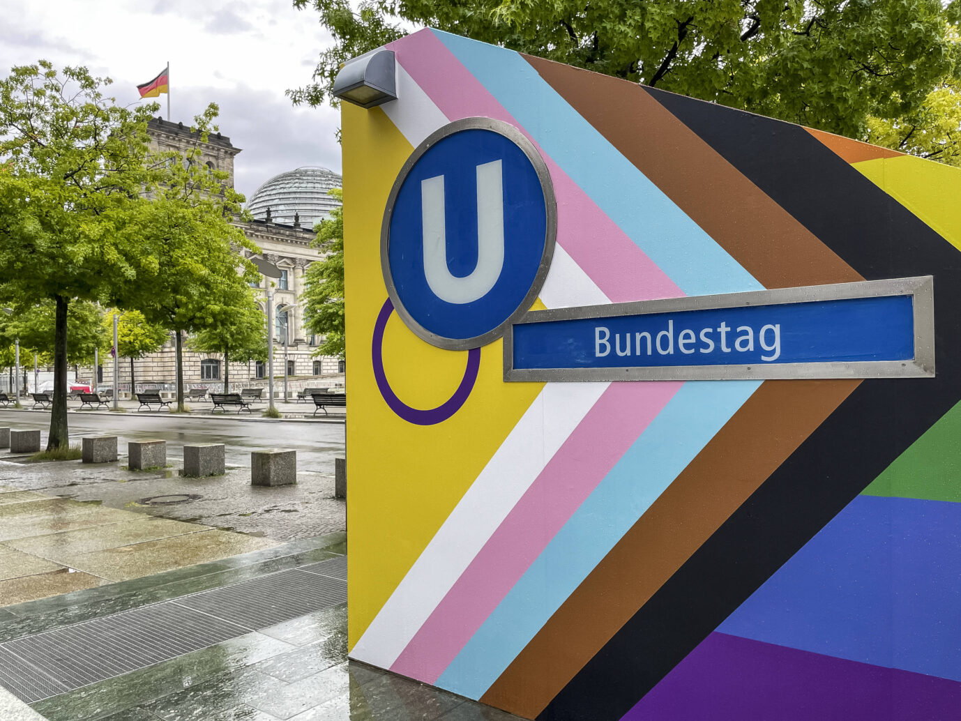 Farbfoto des Berliner U-Bahnausgangs „Bundestag“, beklebt mit den Streifen der dem erweiterten Regenbogen der Progress-Pride-Flagge. Im Hintergrund das Reichstagsgebäude mit deutscher Fahne. Die landeseigene BVG ließ den Eingang zum CSD 2025 entsprechend folieren. Offizielle Beteiligung des Bundestags gab es nicht. Der U-Bahnausgang „Bundestag“ mit Progress-Pride-Folie: Ein Seitenhieb auf die Parlamentspräsidentin?