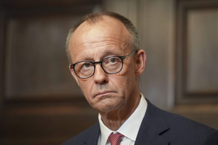 Bundeskanzler Friedrich Merz mit ernstem Gesichtsausdruck. Er trägt eine randlose Brille, einen dunklen Anzug, ein weißes Hemd und eine rot gemusterte Krawatte. Im Hintergrund ist eine dunkle Holzvertäfelung unscharf zu erkennen. Merz blickt direkt in die Kamera, seine Mimik wirkt angespannt und konzentriert.