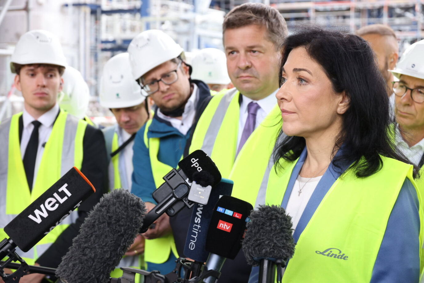 Wirtschaftsministerin Katharina Reiche spricht bei einem Werksbesuch des Gasekonzerns Linde vor Mikrofonen mehrerer Medienvertreter. Sie trägt eine gelbe Warnweste und einen blauen Blazer. Umgeben ist sie von Männern in Schutzhelmen und Sicherheitskleidung, im Hintergrund sind Industrieanlagen zu sehen. Bundeswirtschaftsministerin Katharina Reiche (CDU) besucht ein Chemiewerk: Das Bruttoinlandsprodukt fällt im zweiten Quartal. Foto: IMAGO / Frank Ossenbrink