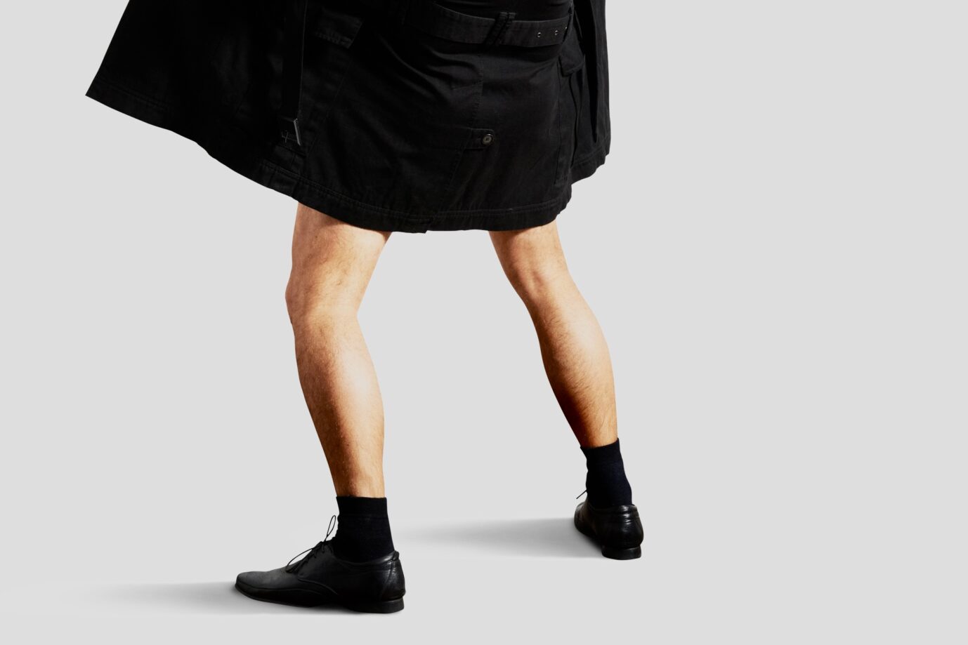 Männerbeine in schwarzen Socken und schwarzen Lederschuhen unter einem kurzen schwarzen Rock, von hinten fotografiert, vor grauem Hintergrund. Mantel auf: Wer im Rechtssinn kein „Mann“ mehr ist, kann straffrei exhibitionieren. Foto: IMAGO / Depositphotos