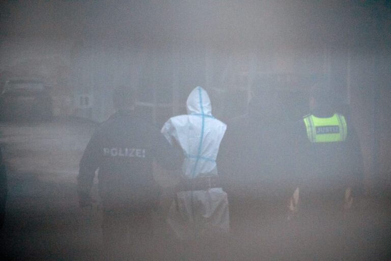 Ein Mann in weißem Schutzanzug mit blauen Streifen wird von mehreren Beamten, darunter einer mit der Aufschrift „POLIZEI“ und einer mit der Warnweste „JUSTIZ“, aus einem Gerichtsgebäude geführt. Die Szene ist durch ein Hindernis unscharf und körnig fotografiert. Hintergrund: Der mutmaßliche Täter des Messerangriffs im Park Schöntal wurde nach der Vorführung beim Haftrichter zurück zum Polizeifahrzeug gebracht. Nach der Bluttat von Aschaffenburg: Der mutmaßliche Täter wird unter Polizeibegleitung aus dem Amtsgericht geführt. Archivbild: picture alliance/dpa | Daniel Vogl