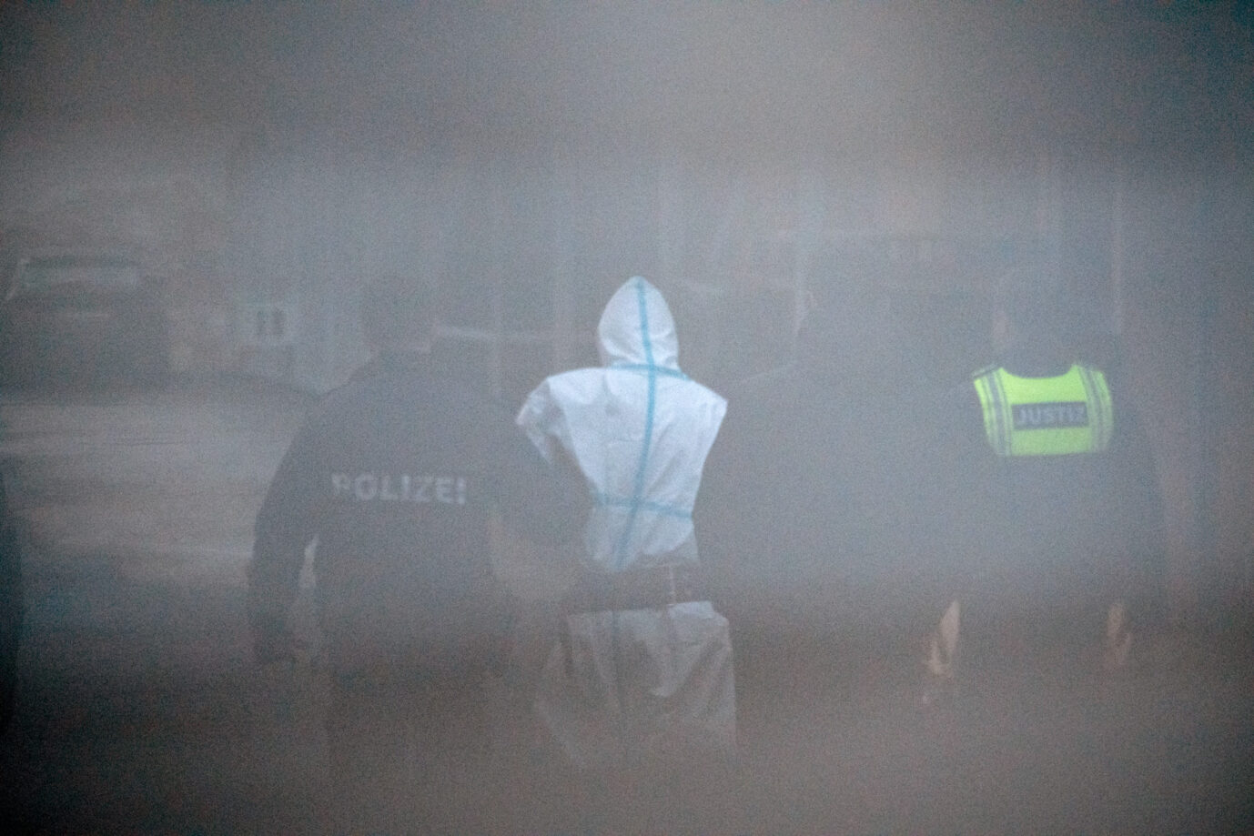 Ein Mann in weißem Schutzanzug mit blauen Streifen wird von mehreren Beamten, darunter einer mit der Aufschrift „POLIZEI“ und einer mit der Warnweste „JUSTIZ“, aus einem Gerichtsgebäude geführt. Die Szene ist durch ein Hindernis unscharf und körnig fotografiert. Hintergrund: Der mutmaßliche Täter des Messerangriffs im Park Schöntal wurde nach der Vorführung beim Haftrichter zurück zum Polizeifahrzeug gebracht. Nach der Bluttat von Aschaffenburg: Der mutmaßliche Täter wird unter Polizeibegleitung aus dem Amtsgericht geführt. Archivbild: picture alliance/dpa | Daniel Vogl