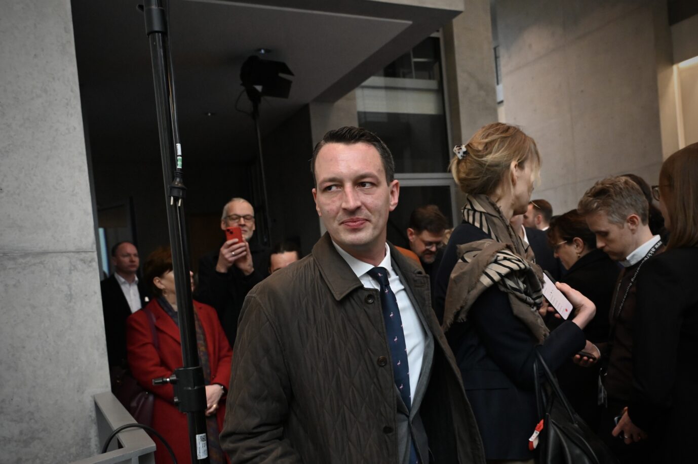 Matthias Helferich, Bundestagsabgeordneter aus Dortmund, steht im Foyer des Reichstagsgebäudes in Berlin. Er trägt einen olivgrünen Steppmantel, weißes Hemd, blauen Schlips mit kleinem Muster und blickt leicht nach rechts. Im Hintergrund sind weitere Personen zu sehen, darunter Abgeordnete und Mitarbeiter, teils in Gespräche vertieft. Eine Frau im Vordergrund hält ein Smartphone mit Notizen. Der Ort wirkt wie eine Zwischenzone vor dem Plenarsaal – mit Betonwänden und gedämpftem Licht. Matthias Helferich im Bundestag: Borussia Dortmund fordert vom Abgeordneten eine Unterlassungserklärung. Foto: picture alliance/dpa | Elisa Schu