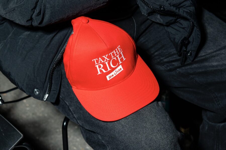 Rote Baseballkappe mit der Aufschrift „Tax the rich“ und dem Logo der Partei Die Linke liegt auf dem Bein einer sitzenden Person, die ein Smartphone in der Hand hält. Marx21 will „Ferat in den Bundestag bringen“ – und hat es geschafft. Der Verfassungsschutz sieht in der Strömung die einzige relevante extremistische Kraft in der Linkspartei. Rote Botschaft: Kappe mit der Aufschrift „Tax the rich“ und Logo der Linkspartei auf der Wahlparty von Ferat Koçak. Foto: picture alliance / Eventpress | Eventpress Jeremy Knowles