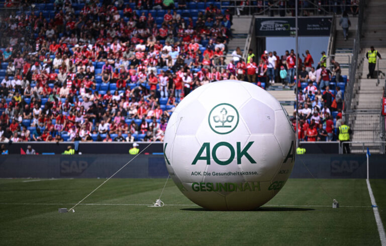 Großer, weißer, aufblasbarer Fußball mit grünem AOK-Logo und dem Schriftzug „AOK. Die Gesundheitskasse“ steht auf dem Rasen eines Fußballstadions. Im Hintergrund sind Zuschauer in rot-weißer Kleidung auf den Tribünen zu sehen, daneben leere Sitzreihen und Ordner in gelben Westen. Das Spielfeld ist für ein Spiel zwischen der TSG 1899 Hoffenheim und dem FC Bayern München vorbereitet. AOK-Werbeball vor einem Fußballspiel zwischen der TSG 1899 Hoffenheim und dem FC Bayern München: Krankenkassen wollen an Werbepraxis festhalten. Foto: IMAGO / Ulmer/Teamfoto