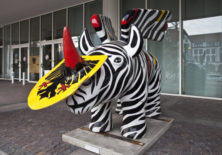 Eine bemalte Nashornfigur mit schwarz-weißen Streifen, roten Flügeln und einem gelben Schild mit dem Dortmunder Stadtwappen steht vor dem Konzerthaus Dortmund. Geflügeltes Nashorn mit Stadtwappen vor dem Dortmunder Konzerthaus: Das Symboltier der Stadt ist frei zugänglich – das Wappen im Wahlkampf dagegen umkämpft. Foto: IMAGO / Zoonar