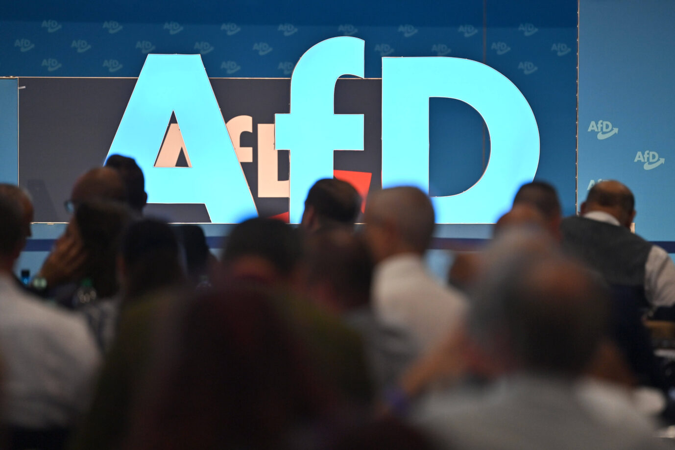 Ein überdimensionales AfD-Logo steht auf einer Bühne. Davor sitzen zahlreiche Menschen mit dem Rücken zur Kamera, teilweise unscharf im Vordergrund. Im Hintergrund ist mehrfach das AfD-Logo auf einer blauen Wand zu sehen. Großes AfD-Logo auf einer Parteiveranstaltung: In Brandenburg gilt die Partei nun als „gesichert rechtsextrem“. Foto: IMAGO / Sven Simon