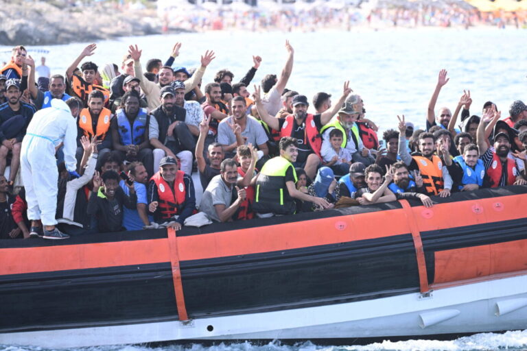Italien wollte Asylverfahren nach Albanien auslagern – der Europäische Gerichtshof stoppt das Modell. Die Richter verlangen Schutz für jede Bevölkerungsgruppe. Homosexualität wird jetzt zum Maßstab für sichere Herkunftsstaaten. Ein überfülltes Schlauchboot mit Dutzenden Migranten auf dem Mittelmeer. Die meisten tragen Rettungswesten und winken oder zeigen Handzeichen in Richtung Kamera. Im Vordergrund ist ein Mann in weißem Schutzanzug zu sehen, der offenbar unterstützt. Im Hintergrund ist eine felsige Küste mit verschwommenem Strandbereich zu erkennen. Das Bild symbolisiert eine typische Ankunftsszene im Rahmen der irregulären Migration nach Europa. Migranten freuen sich bei der Ankunft in Europa: Der Europäische Gerichtshof schränkt die Auswahl sicherer Herkunftsländer massiv ein. Foto: IMAGO / ZUMA Press