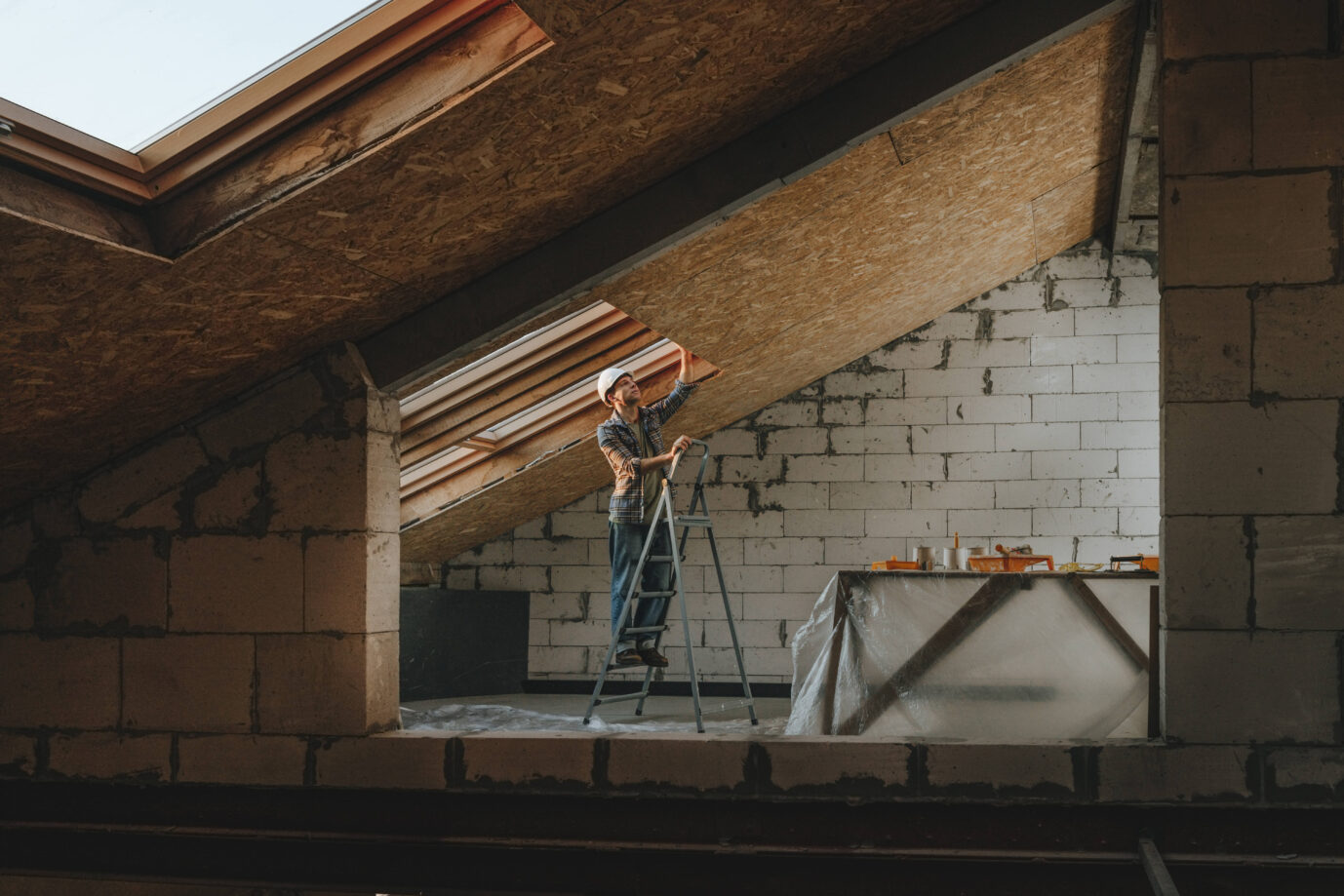 Ein junger Mann hämmert hier zwar nicht, aber so ist das Wort im Alltext – mit Bauhelm steht auf einer Leiter und arbeitet an einer Dachschräge in einem teilsanierten Altbau. Die Wände bestehen aus unverputzten Porenbetonsteinen, auf einem Tisch im Hintergrund liegen Werkzeuge. Tageslicht fällt durch mehrere Dachfenster. Dachausbau unter Eigenregie: In vielen alten Häusern warten Überraschungen hinter jeder Wand – und unter jedem Ziegel. Foto: IMAGO / Westend61