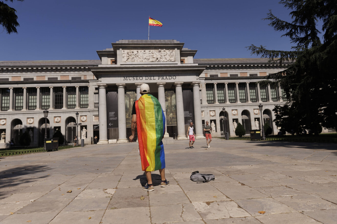 Vor einem antik anmutenden Museum – es handelt sich um das Museo del Prado in Madrid – läuft ein Mann mit einer umgehängten Regenbogenflagge – Symbolbild für den woken Totalitarismus