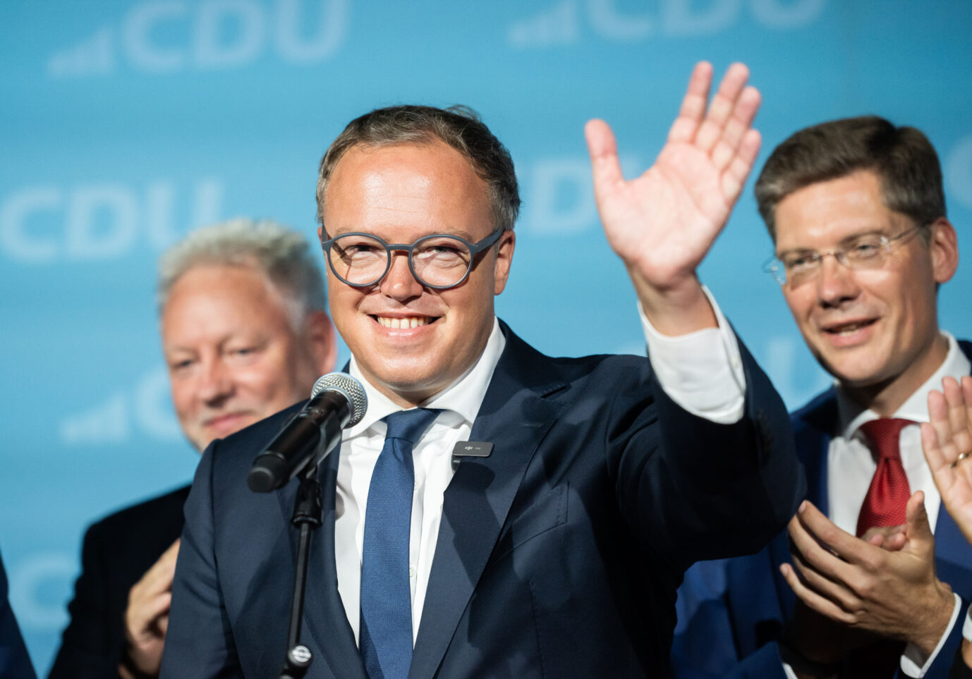 Ein Mann im Anzug und mit Brille winkt lächelnd in die Menge, während er an einem Rednerpult mit Mikrofon steht. Neben ihm applaudieren weitere Personen. Thüringens Ministerpräsident Mario Voigt (CDU) feiert nach der Landtagswahl: Die Plagiatsvorwürfe stehen weiterhin im Raum. Foto: IMAGO / Funke Foto Services