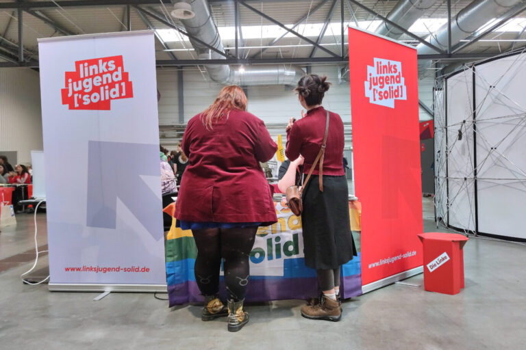 Zwei Personen stehen mit dem Rücken zur Kamera an einem Infostand der Jugendorganisation „Linksjugend [’solid]“. Links und rechts des Tisches sind große Banner mit dem roten Logo der Gruppe aufgestellt. Der Tisch ist mit einer Regenbogenfahne behangen, darauf liegt Informationsmaterial. Die Veranstaltung findet in einer großen Messehalle mit sichtbaren Lüftungsrohren und Betonboden statt. Auf einem kleinen roten Würfel neben dem Stand ist das Logo der Partei „Die Linke“ zu sehen. Im Hintergrund sind weitere Besucher und Stände erkennbar. Die Linksjugend „Solid“ auf dem Parteitag der Linken in Halle (Symbolbild): Die Staatsanwaltschaft prüft einen antisemitischen Tweet. Foto: IMAGO / dts Nachrichtenagentur
