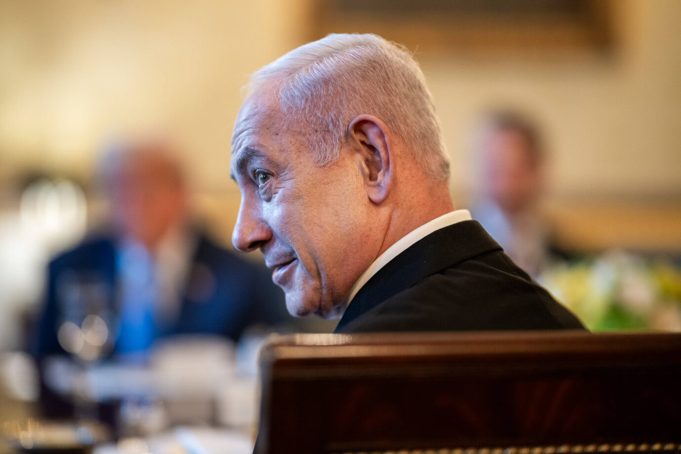 Der israelische Ministerpräsident Benjamin Netanyahu sitzt in einem holzvertäfelten Stuhl, leicht im Profil nach links gewandt. Er trägt einen dunklen Anzug und ein weißes Hemd. Im unscharfen Hintergrund sitzen weitere Personen an einem Tisch, darunter US-Präsident Donald Trump. Vor Netanyahu stehen Gläser und Geschirr, die Szene spielt am 7. Juli 2025 im Blue Room des Weißen Hauses bei einem bilateralen Abendessen. ﻿﻿Benjamin Netanyahu im Weißen Haus: Er will die Hamas „vollständig besiegen“. Foto: IMAGO / ZUMA Press Wire