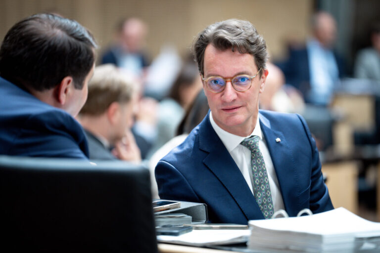 Ein Mann im blauen Anzug mit gemusterter Krawatte und Brille sitzt in einem Parlament oder Sitzungssaal und schaut zur Seite. Vor ihm liegen Aktenstapel, ein Smartphone und ein Ringbuch. Im Hintergrund sitzen weitere Personen, teils unscharf im Gespräch vertieft. Nordrhein-Westfalens Ministerpräsident Hendrik Wüst (CDU) im Bundesrat: Rechnungsprüfer kritisieren den Haushalt seiner schwarz-grünen Regierung. Foto: IMAGO / Political-Moments