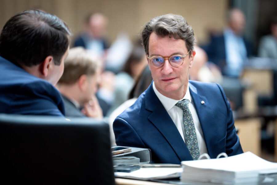 Ein Mann im blauen Anzug mit gemusterter Krawatte und Brille sitzt in einem Parlament oder Sitzungssaal und schaut zur Seite. Vor ihm liegen Aktenstapel, ein Smartphone und ein Ringbuch. Im Hintergrund sitzen weitere Personen, teils unscharf im Gespräch vertieft. Nordrhein-Westfalens Ministerpräsident Hendrik Wüst (CDU) im Bundesrat: Rechnungsprüfer kritisieren den Haushalt seiner schwarz-grünen Regierung. Foto: IMAGO / Political-Moments