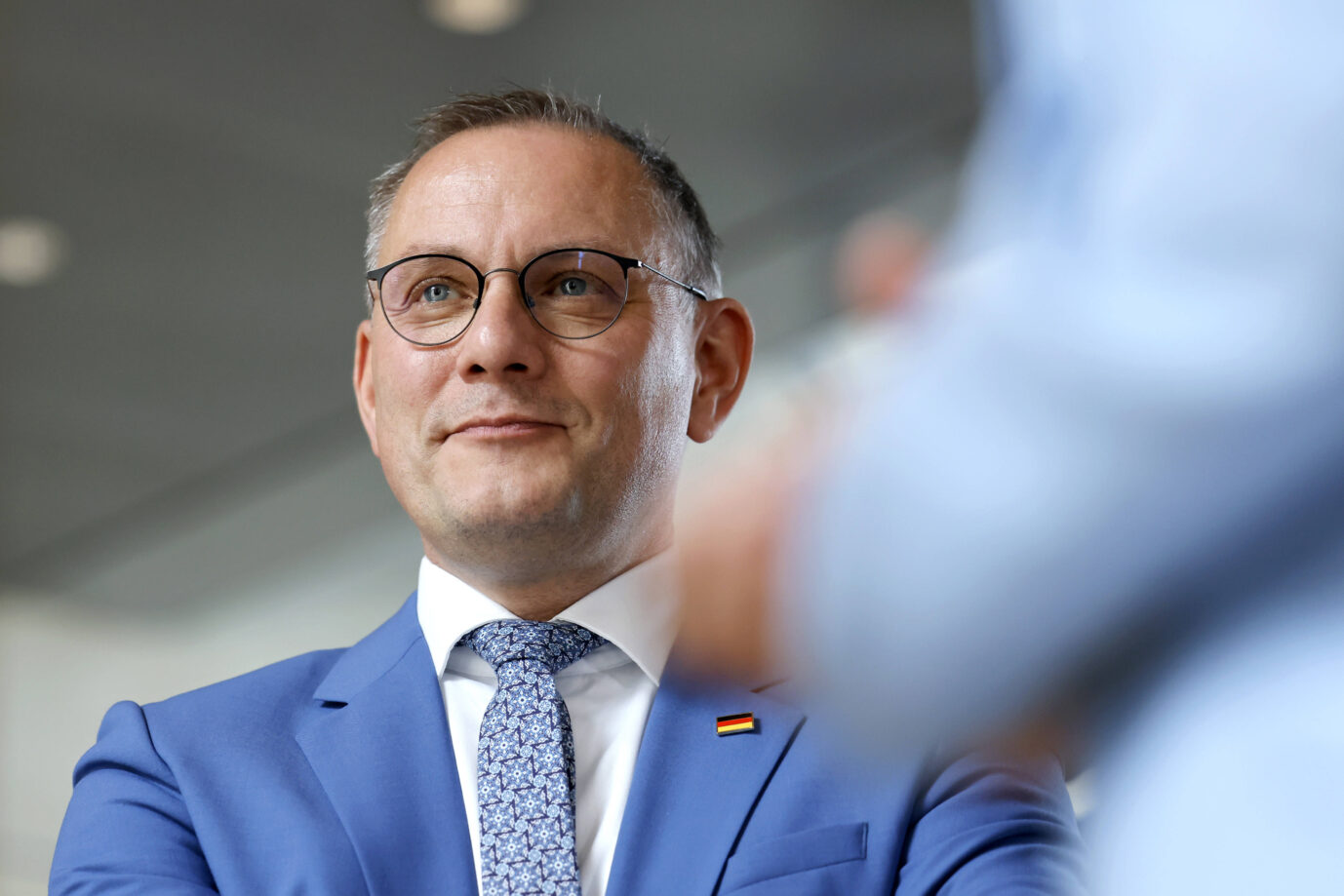 Ein Mann mit Brille, blauem Anzug, weißem Hemd und gemusterter Krawatte steht in einem Innenraum und blickt leicht lächelnd zur Seite. Am Revers trägt er eine Anstecknadel in den Farben der deutschen Flagge. Tino Chrupalla: Der AfD-Chef wirft Israel im Gaza-Krieg „Verbrechen“ vor und fordert politischen Druck. Foto: IMAGO / Panama Pictures