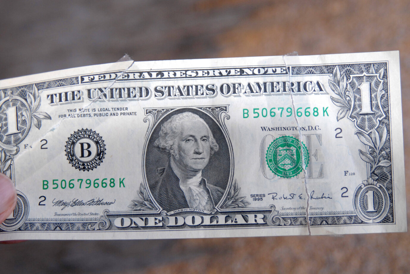 Ein stark abgenutzter und an mehreren Stellen eingerissener US-Ein-Dollar-Schein der Serie von 1995. In der Mitte ist das Porträt von George Washington zu sehen. Der Geldschein trägt die Seriennummer B50679668K und stammt aus dem Federal Reserve District New York („B“). Die Unterschriften unten links und rechts stammen von Mary Ellen Withrow (Treasurer of the United States) und Robert E. Rubin (Secretary of the Treasury). Das Papier ist vergilbt und weist deutliche Faltspuren auf. Der Schein wird von einer Hand am linken Rand gehalten, im Hintergrund ist eine unscharfe Stein- oder Asphaltfläche zu erkennen. Ein eingerissener US-Ein-Dollar-Schein: Symbol einer schwächelnden Währung. Foto: IMAGO / Dean Pictures