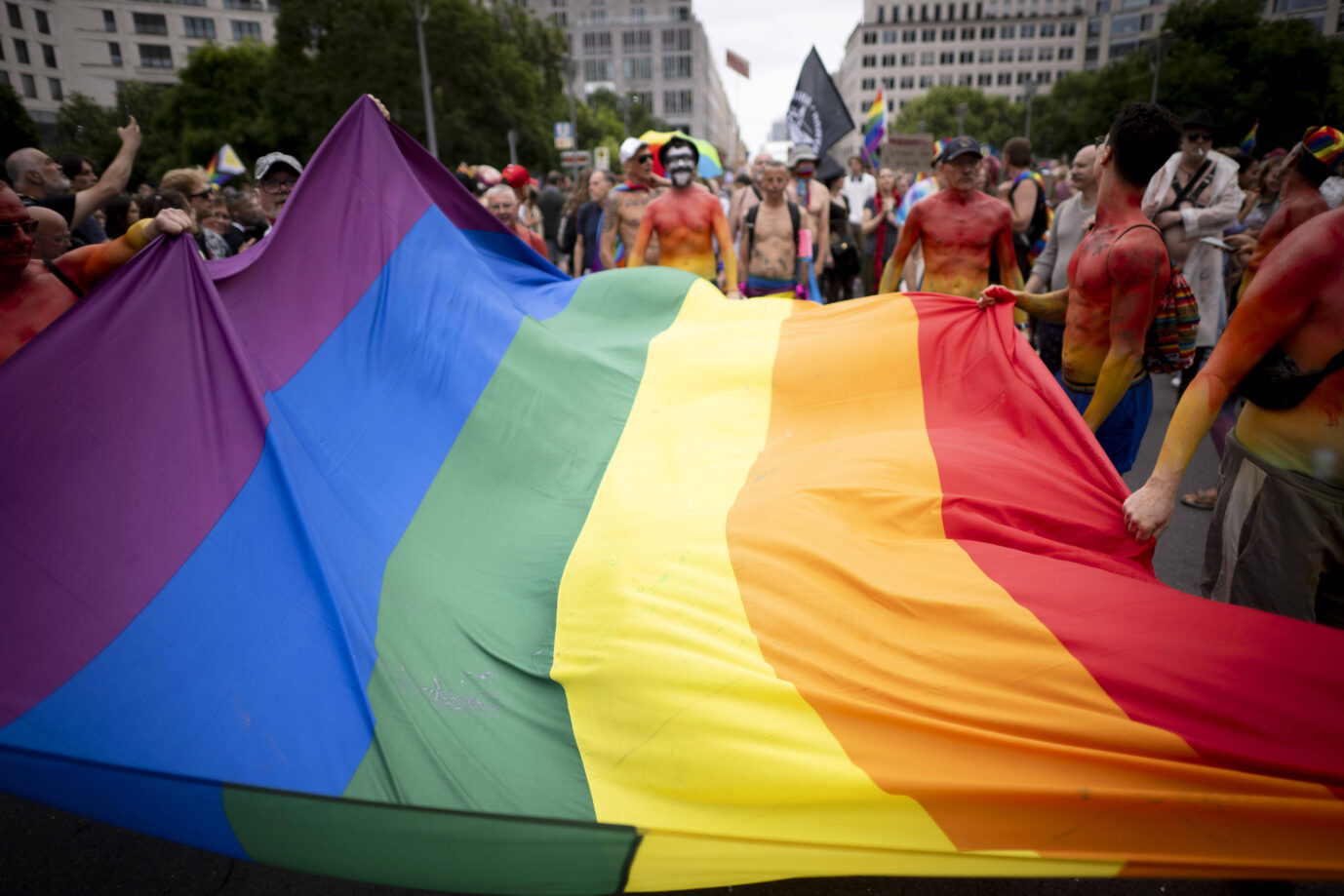 Teilnehmer des Christopher Street Day in Berlin halten gemeinsam eine große Regenbogenfahne. Im Hintergrund stehen bunt bemalte Menschen, viele mit nacktem Oberkörper, einige mit Flaggen oder Transparenten. Die Szene spielt sich auf einer gesperrten Straße zwischen Hochhäusern ab. Bunt angemalte Männer tragen eine Regenbogenflagge: Auch der Bundestag war beim CSD dabei. Foto: IMAGO / IPON