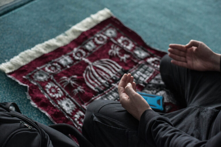 Hände eines betenden Mannes über einem roten Gebetsteppich mit Muster, daneben eine Tasche. Symbolbild zum neuen Vertrag in Schleswig-Holstein, der Muslimen Freistellung an Ramadan- und Opferfest ermöglicht. Ein Mann betet auf einem Gebetsteppich: Schleswig-Holstein erkennt künftig Ramadan- und Opferfest als islamische Feiertage an. Foto: picture alliance/dpa | Eman Helal