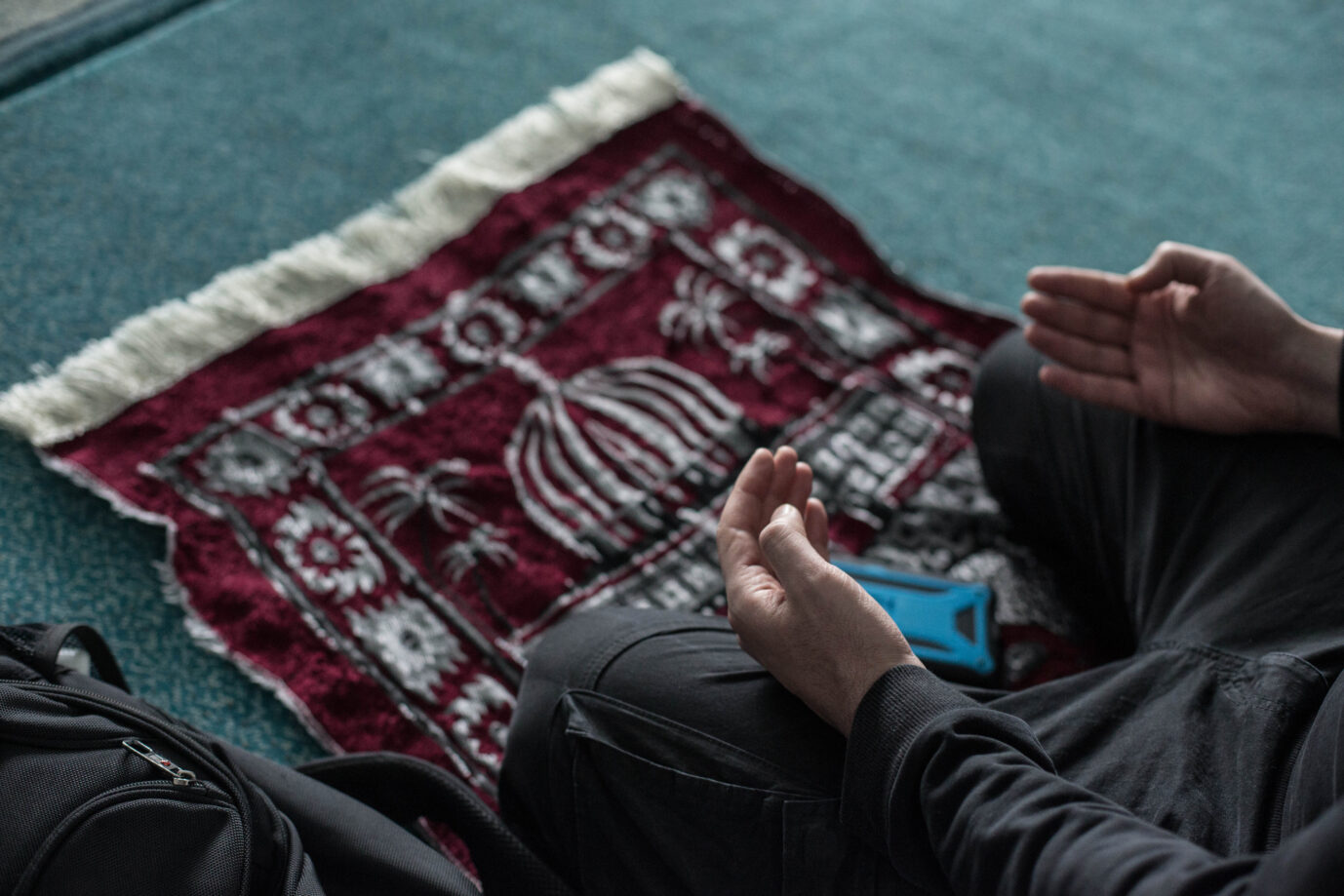 Hände eines betenden Mannes über einem roten Gebetsteppich mit Muster, daneben eine Tasche. Symbolbild zum neuen Vertrag in Schleswig-Holstein, der Muslimen Freistellung an Ramadan- und Opferfest ermöglicht. Ein Mann betet auf einem Gebetsteppich: Schleswig-Holstein erkennt künftig Ramadan- und Opferfest als islamische Feiertage an. Foto: picture alliance/dpa | Eman Helal