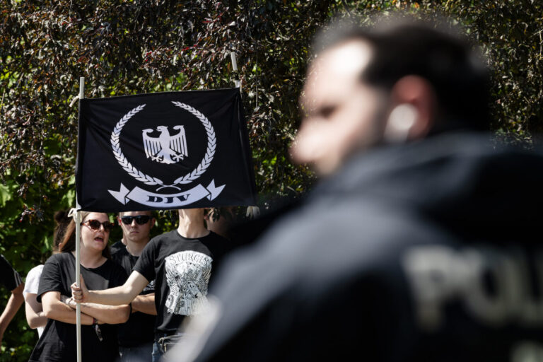 Mehrere Menschen in schwarzer Kleidung halten eine schwarze Fahne mit Adler-Emblem und der Aufschrift „DJV“, im Vordergrund unscharf ein Polizist. Die Aufnahme entstand bei einer Demonstration der rechtsextremen Gruppe Deutsche Jugend voran gegen die Marzahn Pride in Berlin. Anhänger der „Deutschen Jugend voran“ in Berlin: Polizei ermittelt wegen Angriffen auf Journalisten. Foto: picture alliance / Eventpress | EVENTPRESS Jeremy Knowles
