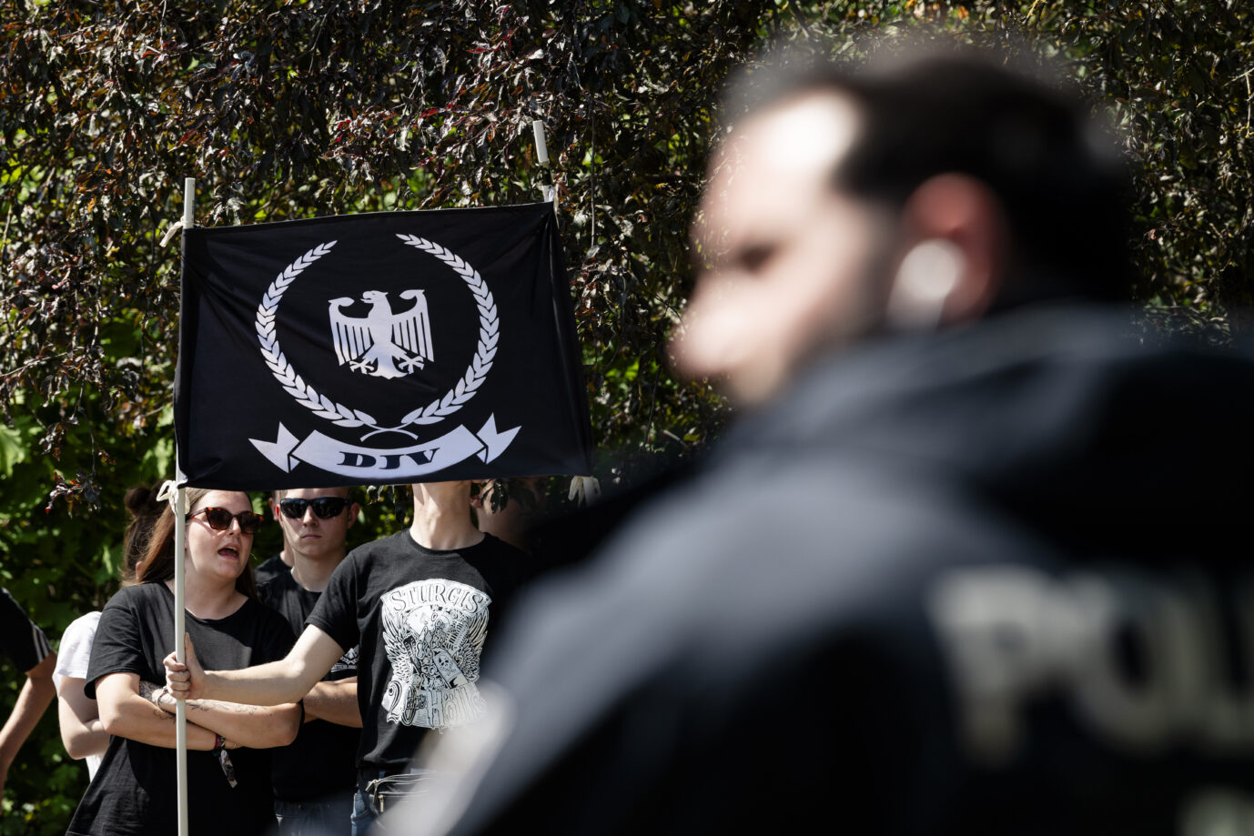 Mehrere Menschen in schwarzer Kleidung halten eine schwarze Fahne mit Adler-Emblem und der Aufschrift „DJV“, im Vordergrund unscharf ein Polizist. Die Aufnahme entstand bei einer Demonstration der rechtsextremen Gruppe Deutsche Jugend voran gegen die Marzahn Pride in Berlin. Anhänger der „Deutschen Jugend voran“ in Berlin: Polizei ermittelt wegen Angriffen auf Journalisten. Foto: picture alliance / Eventpress | EVENTPRESS Jeremy Knowles