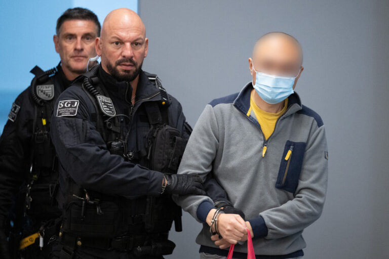 Ein Justizbeamter führt einen gefesselten Mann mit Mundschutz in den Gerichtssaal. Der Mann ist Jian G., früherer Mitarbeiter von AfD-Politiker Maximilian Krah, der vor dem Oberlandesgericht Dresden wegen Spionage für China angeklagt ist. Justizbeamte führen Jian G. in den Gerichtssaal: Der frühere Krah-Mitarbeiter ist in Dresden wegen Spionage verurteilt worden. Foto: picture alliance/dpa | Sebastian Kahnert