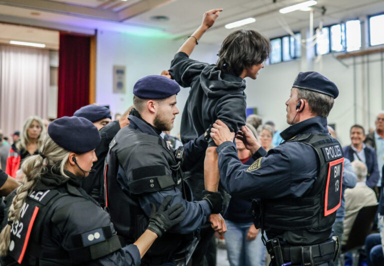 Polizeibeamte in Schutzkleidung führen einen jungen Mann aus der Hermann-Hepper-Halle in Tübingen. Der Mann trägt einen schwarzen Pullover mit Antifa-Logo und hatte zusammen mit anderen Demonstranten die Diskussionsrunde von Oberbürgermeister Boris Palmer und AfD-Landeschef Markus Frohnmaier gestört. Störung der Tübinger Debatte zwischen Palmer und Frohnmaier: Antifa-Pullover auf dem Weg nach draußen. Foto: picture alliance/dpa | Christoph Schmidt