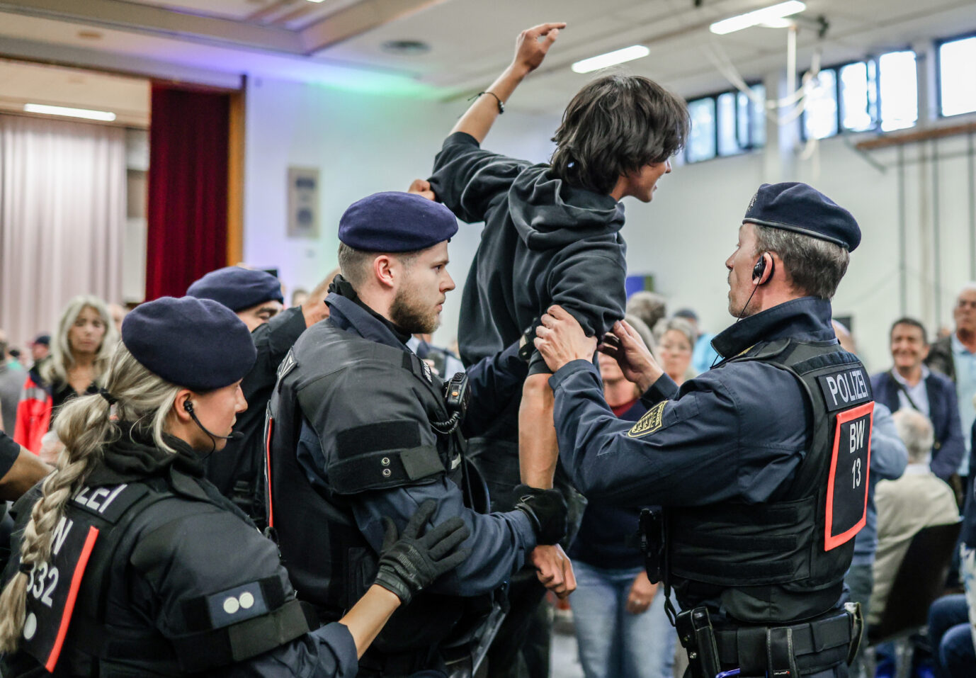 Polizeibeamte in Schutzkleidung führen einen jungen Mann aus der Hermann-Hepper-Halle in Tübingen. Der Mann trägt einen schwarzen Pullover mit Antifa-Logo und hatte zusammen mit anderen Demonstranten die Diskussionsrunde von Oberbürgermeister Boris Palmer und AfD-Landeschef Markus Frohnmaier gestört. Störung der Tübinger Debatte zwischen Palmer und Frohnmaier: Antifa-Pullover auf dem Weg nach draußen. Foto: picture alliance/dpa | Christoph Schmidt
