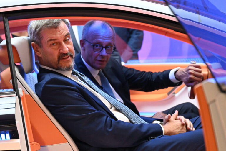 Zwei Männer in dunklen Anzügen sitzen nebeneinander in einem orange-weißen Fahrzeug mit futuristisch gestaltetem Innenraum. Der Mann links blickt nach außen, der Mann rechts greift an das Lenkrad. Beide befinden sich in einem Ausstellungsfahrzeug auf der Automobilmesse IAA in München. Sitzprobe auf der IAA: Bundeskanzler Friedrich Merz am Steuer neben Bayerns Ministerpräsident Markus Söder im Messefahrzeug. Foto: picture alliance/dpa | Sven Hoppe