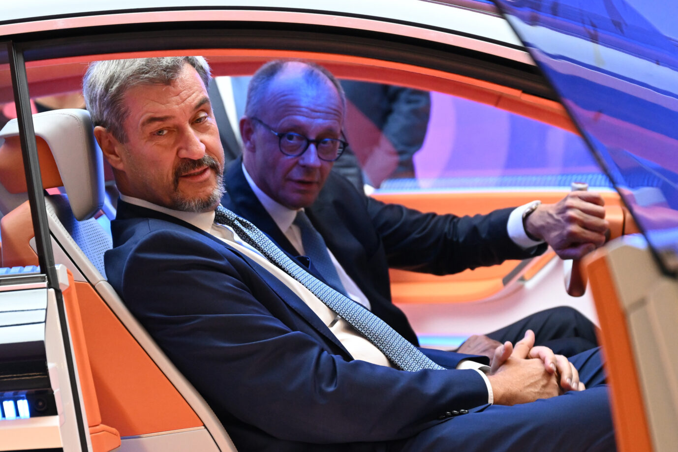 Zwei Männer in dunklen Anzügen sitzen nebeneinander in einem orange-weißen Fahrzeug mit futuristisch gestaltetem Innenraum. Der Mann links blickt nach außen, der Mann rechts greift an das Lenkrad. Beide befinden sich in einem Ausstellungsfahrzeug auf der Automobilmesse IAA in München. Sitzprobe auf der IAA: Bundeskanzler Friedrich Merz am Steuer neben Bayerns Ministerpräsident Markus Söder im Messefahrzeug. Foto: picture alliance/dpa | Sven Hoppe