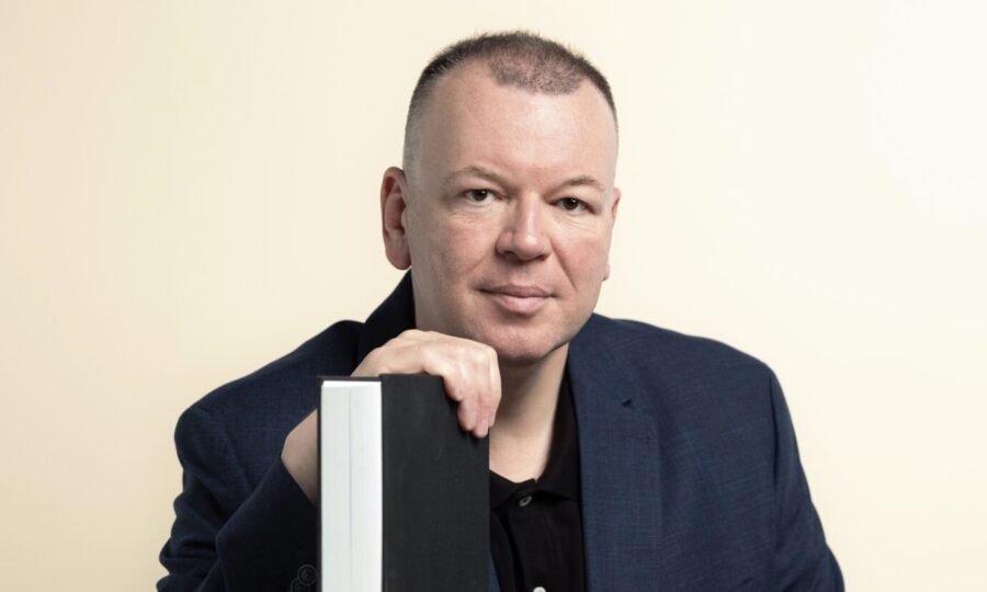 Stefan Weber, Kommunikationswissenschaftler aus Wien, sitzt an einem Tisch und lehnt sich mit der Hand an ein aufgestelltes Buch. Er trägt dunkles Sakko und schwarzes Hemd und blickt in die Kamera. Stefan Weber: Der Wiener Kommunikationswissenschaftler und Plagiatsprüfer warnt vor Kaufholds Konzept einer „Systemaufsicht“. Foto: Joachim Bergauer