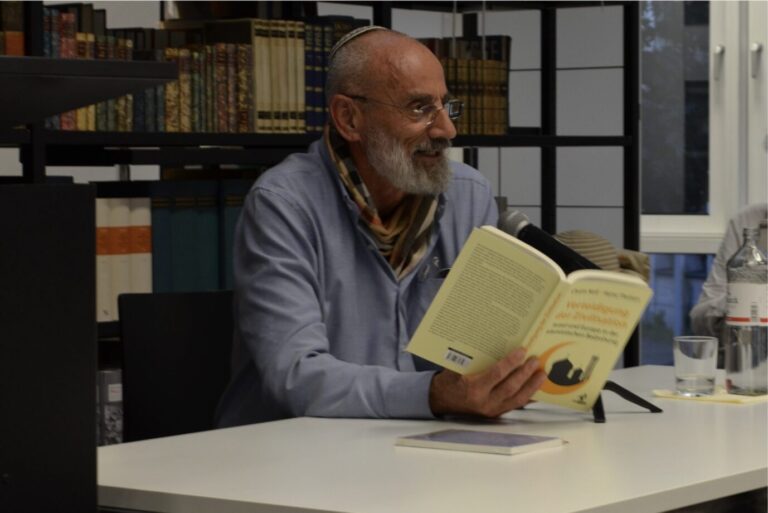 Der israelische Autor Chaim Noll sitzt vor einer Bücherwand, hält sein Buch in der Hand und liest daraus
