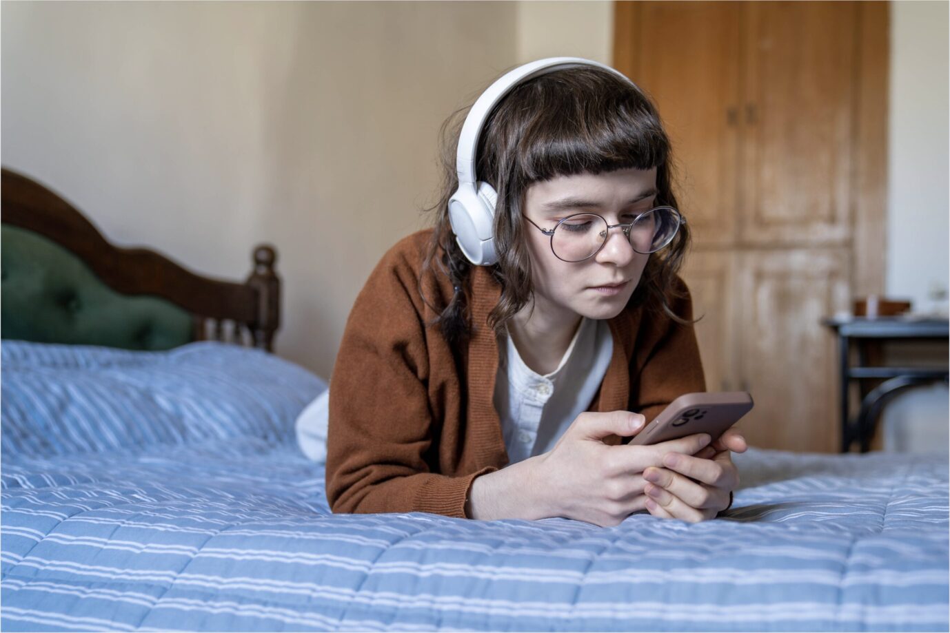 Auf einem mit einem mit einem blauen Bettlaken bezogenen Bett liegt ein junges Mädchen mit Problempony und nutzt ihr Smartphone – vermutlich nutzt sie gerade soziale Medien – und ist psychisch krank, denn durch die sozialen Medien werden die kiddies krank