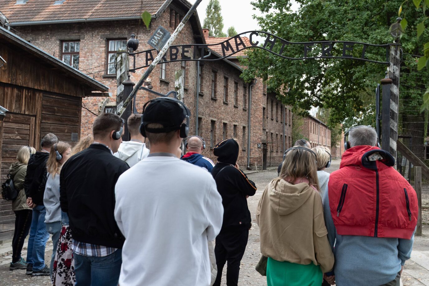 Eine Gruppe von Menschen mit Kopfhörern steht vor dem Tor mit der Aufschrift „Arbeit macht frei“ in der Gedenkstätte Auschwitz. Die Aufnahme zeigt Besucher einer Führung durch das ehemalige Konzentrationslager, das im Zentrum der aktuellen Debatte um Holocaust-Bildung an Schulen steht. Besuchergruppe am Eingangstor der Gedenkstätte Auschwitz: Vertreter fordern, daß Schüler früher mit der Aufarbeitung der NS-Zeit und des Holocaustes beginnen. Foto: IMAGO / Revierfoto